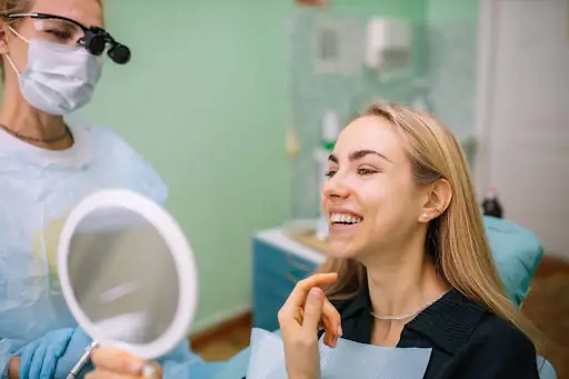 A woman looks into a mirror and smiles at a dental office. A densist stands next to the woman wearing scrubs, a facemask, and dental loupes.]